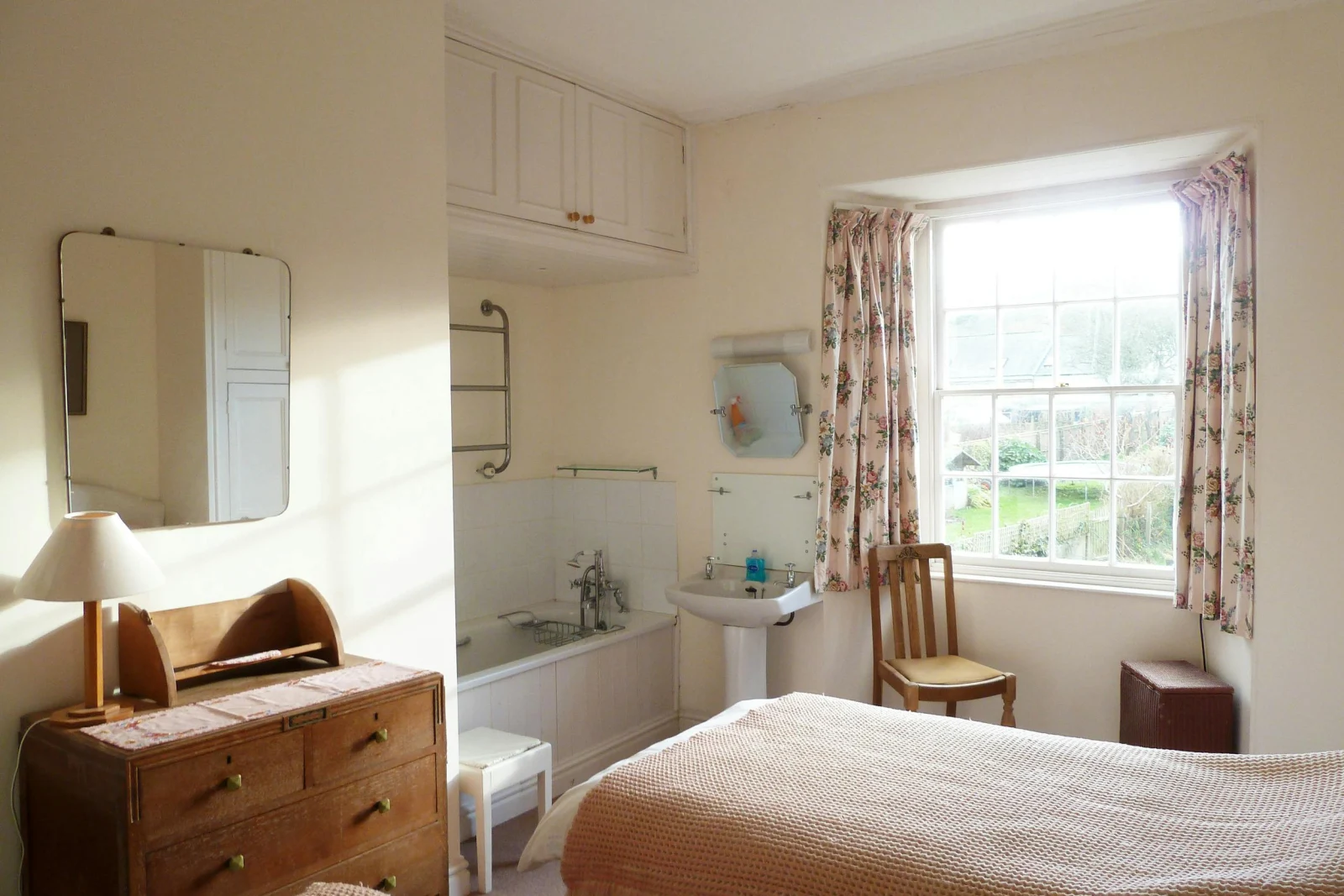 Twin single beds, chest of drawers with a lamp and mirror above, chair, bath and wash hand basin