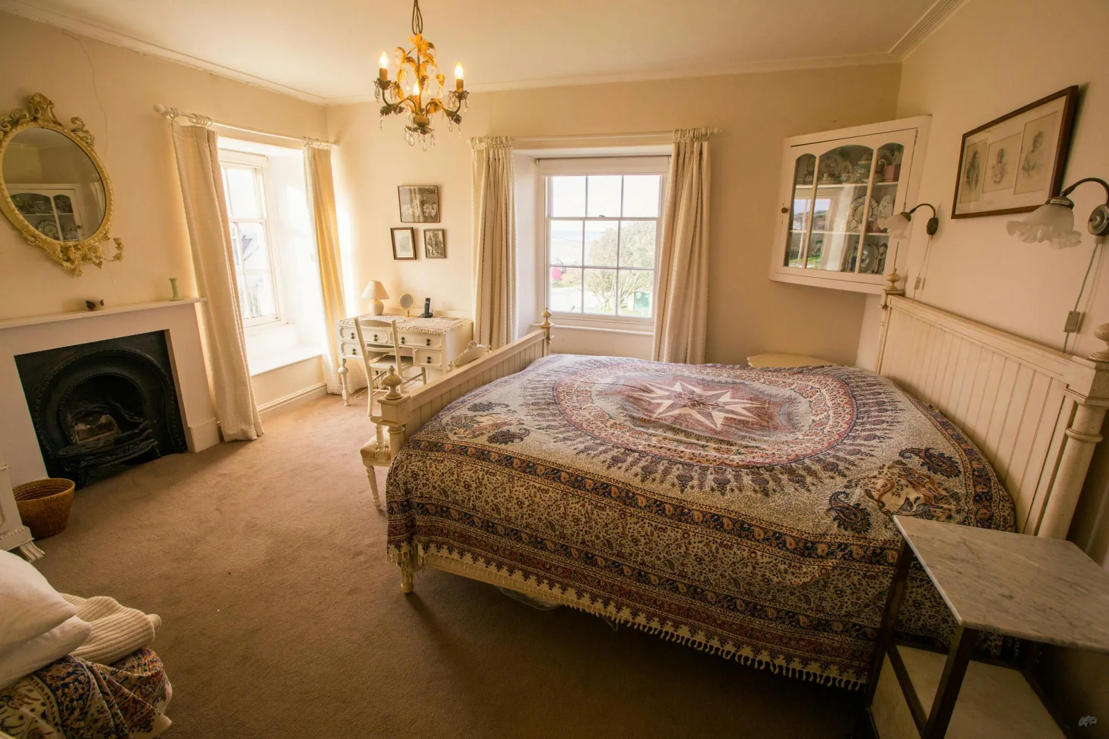 Bedroom with open fireplace, bed with bedside tables, dressing table with a lamp and chair, two windows