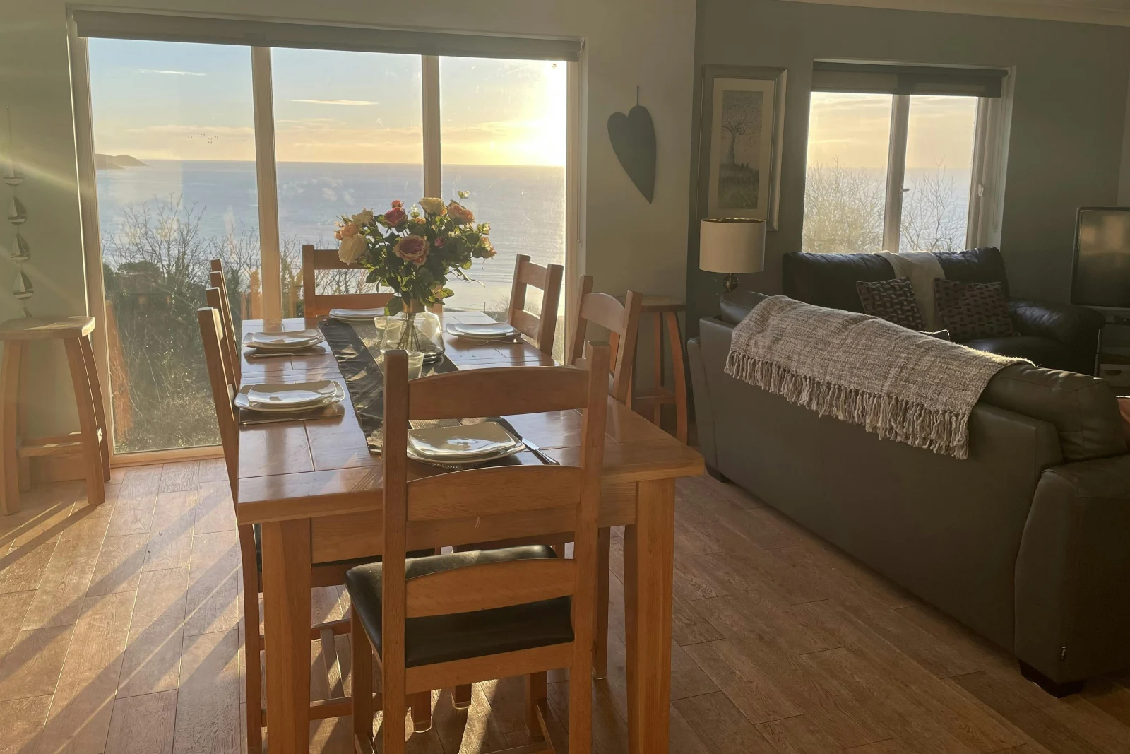 Dining table with six chairs set for breakfast with flowers in vase; back of sofa leading in to lounge