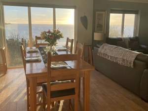 Dining table with six chairs set for breakfast with flowers in vase; back of sofa leading in to lounge