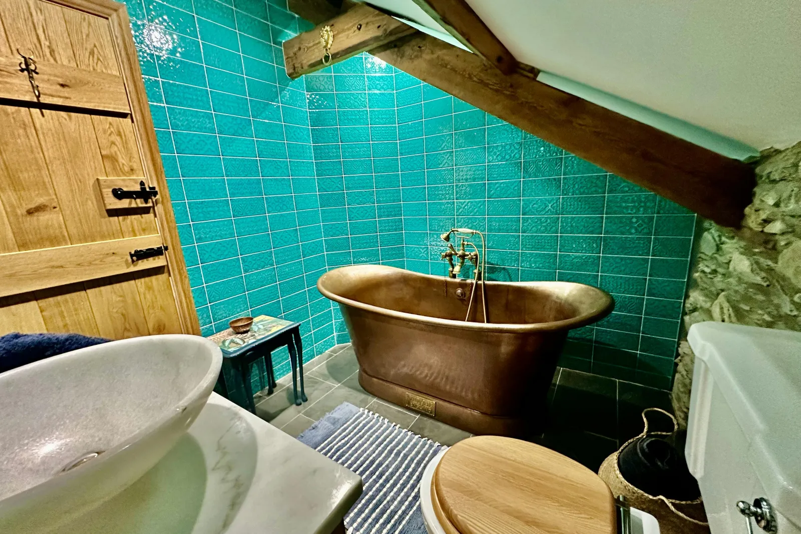 copper bath, toilet with wooden lid, hand wash basin on a marble worktop. Blue wall tiles.